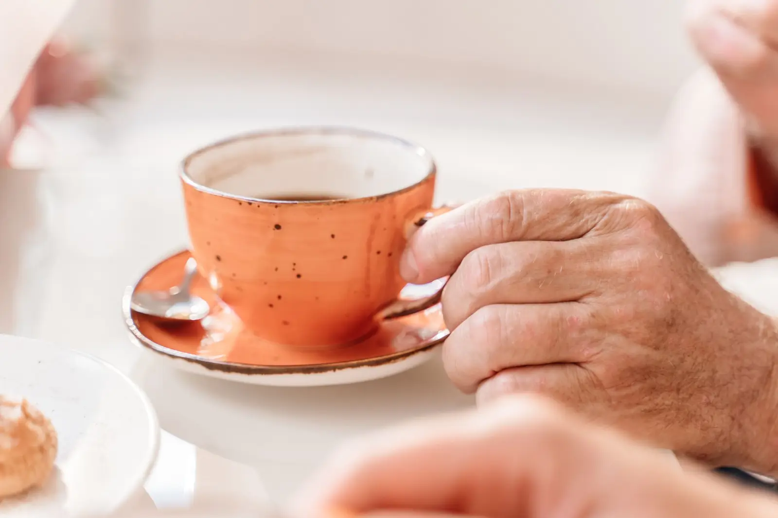 An older person's hand holding a speckled orange coffee cup on its saucer
