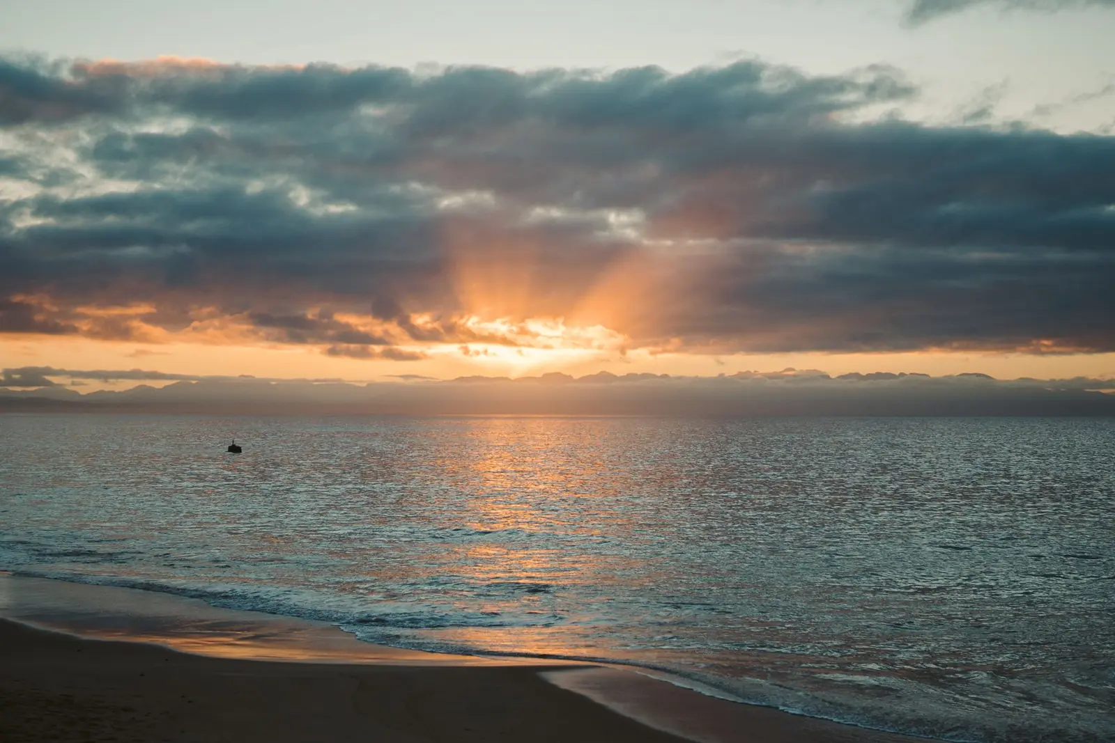 Gentle waves reaching a beach as the sun rises through clouds over the ocean