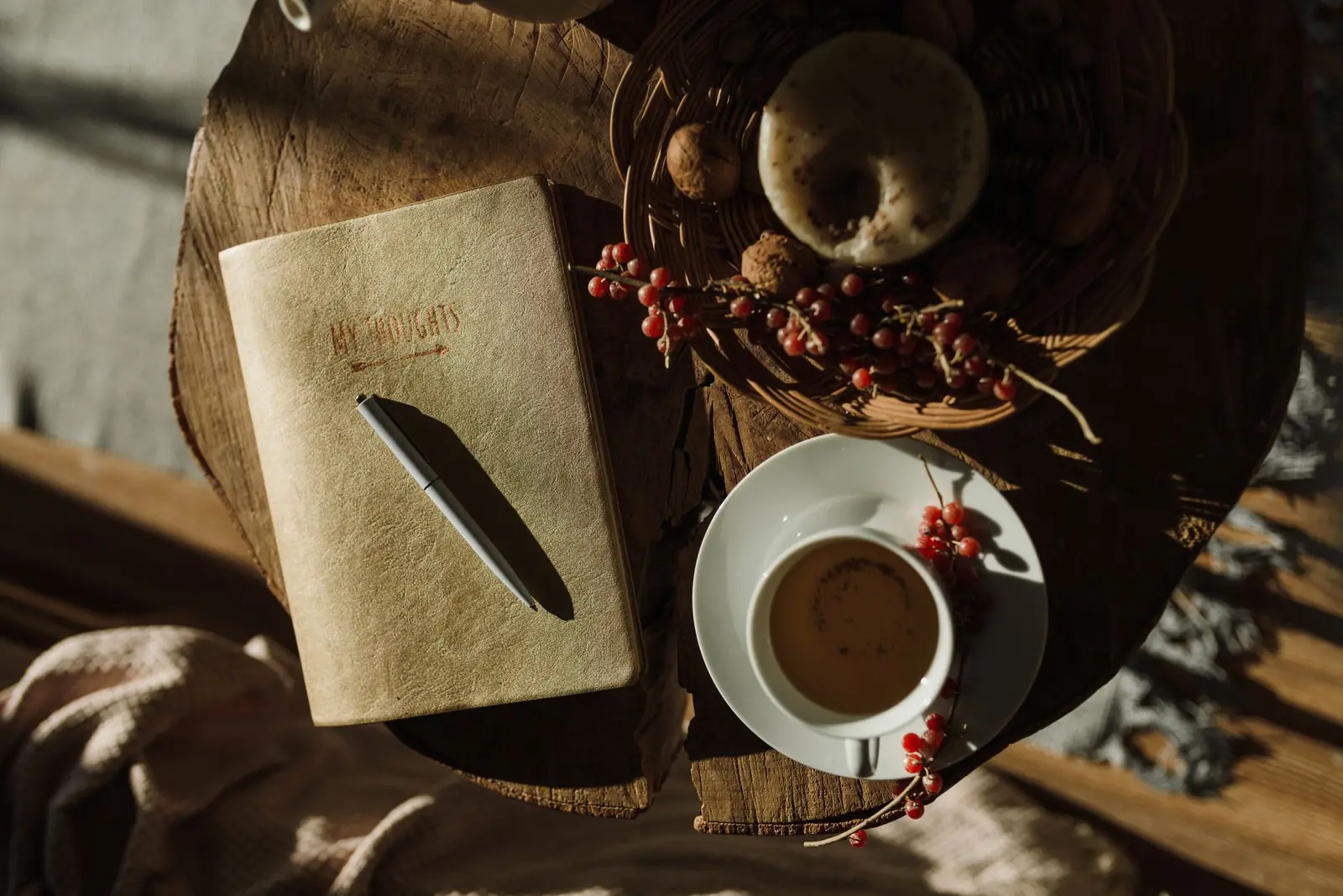 An overhead view of a gold notebook with a pen, a cup of tea on a white saucer and sprigs of red berries on a sunlit wooden table