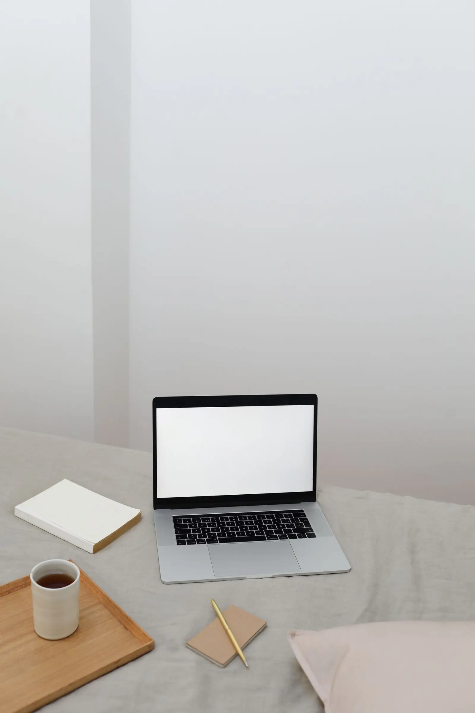 A laptop, notebook and cup of tea on a bed in soft daylight.