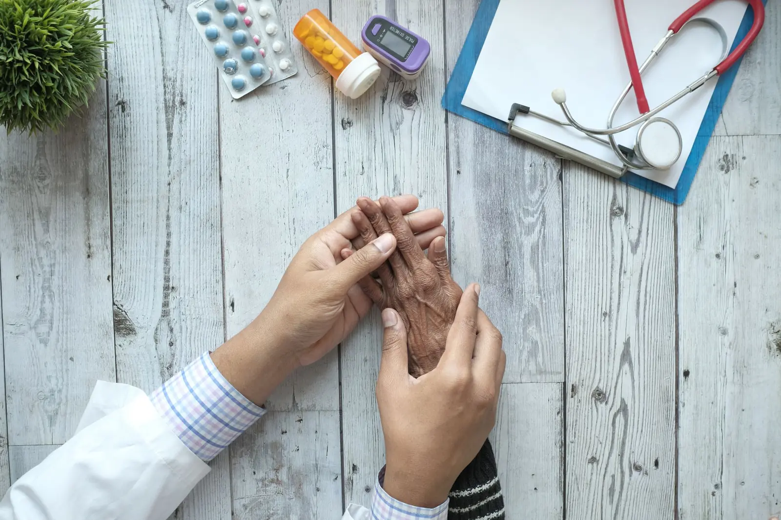 A doctor's hands gently holding an older patient's hand on a desk beside a stethoscope