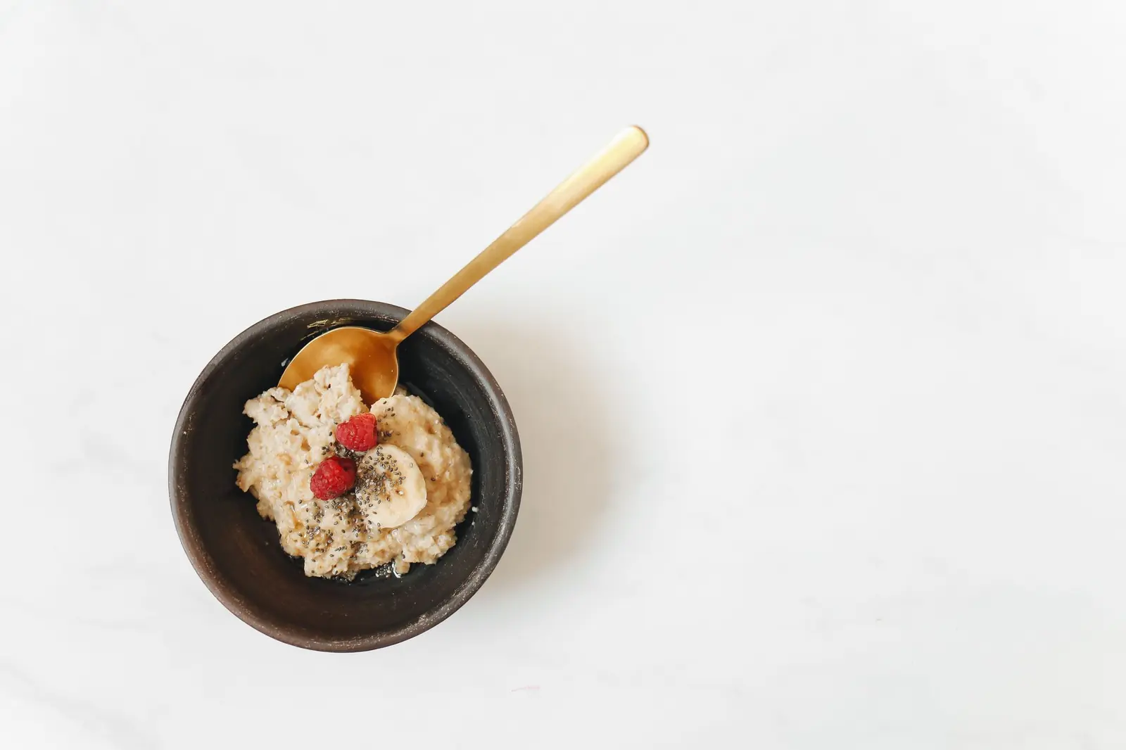 A bowl of oatmeal topped with raspberries, banana and chia seeds on a white marble surface