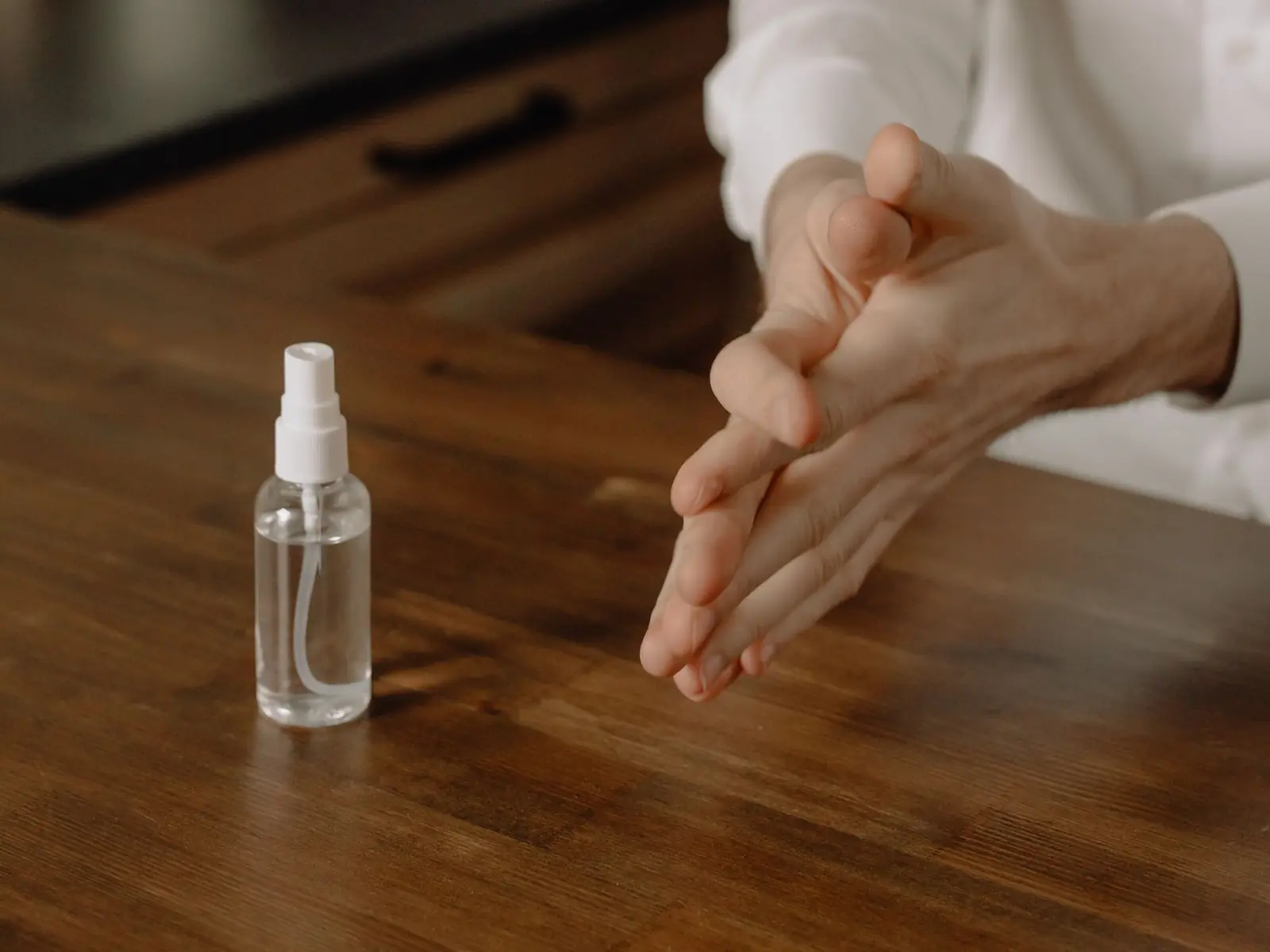 Hands rubbing in sanitizer beside a small clear spray bottle on a wooden table