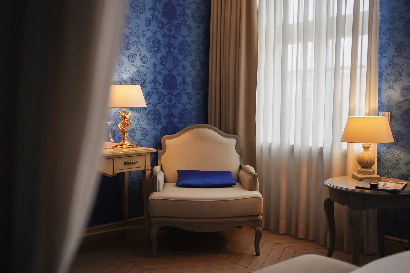An empty upholstered armchair by a curtained window, with two lit table lamps on either side