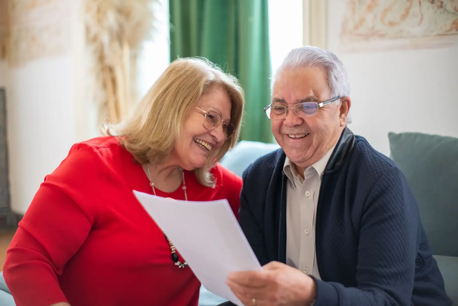 An older couple smiling as they read a document together on a living room sofa