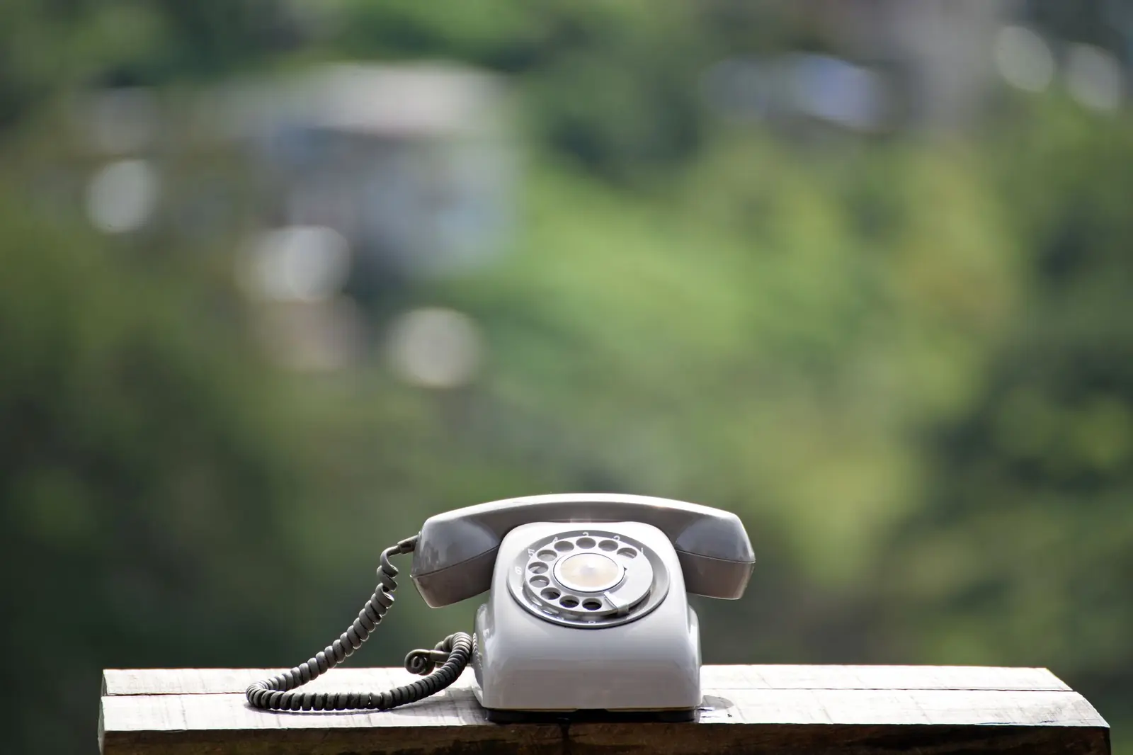 A gray rotary telephone on a wooden ledge outdoors against a soft green background