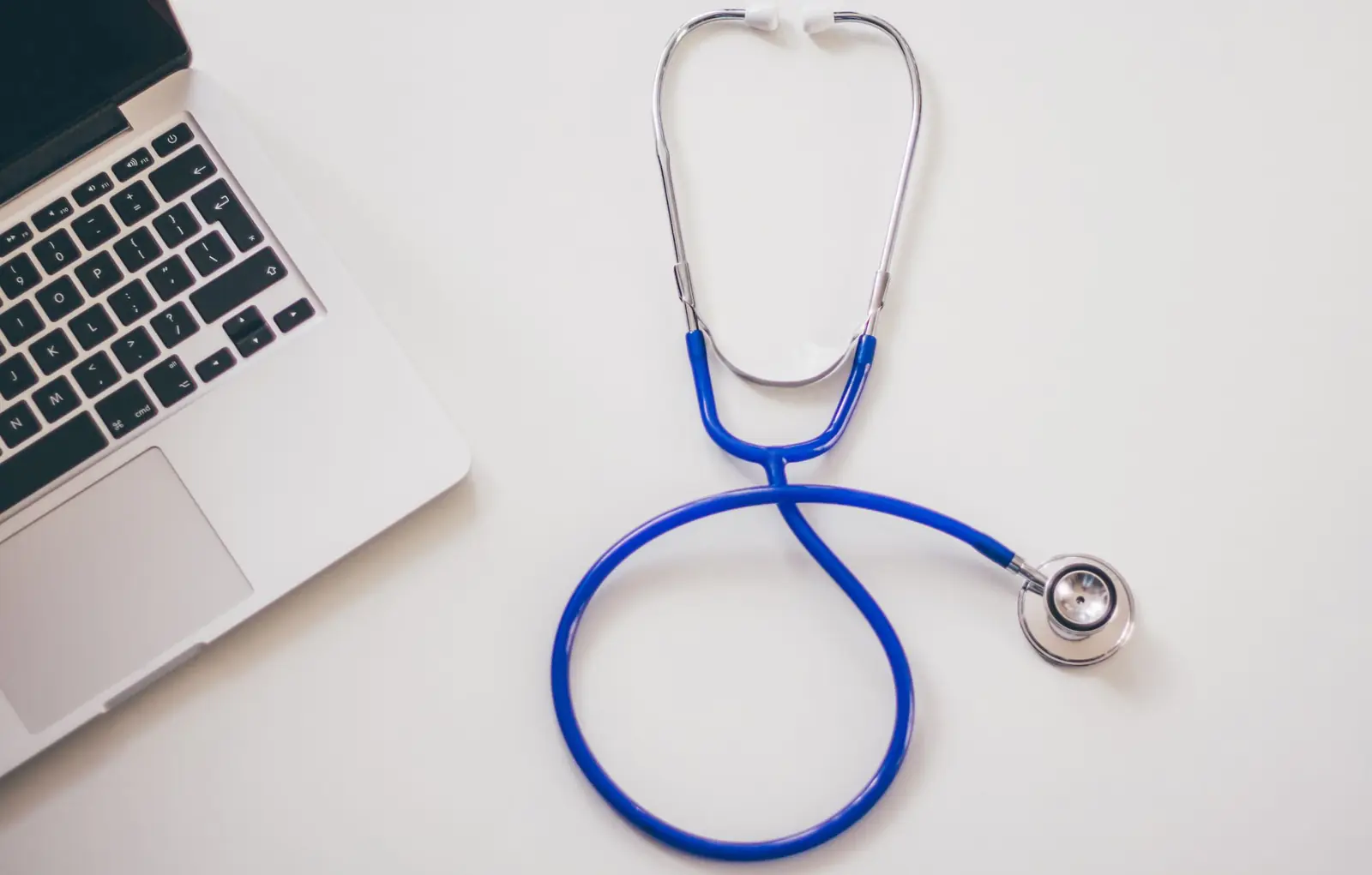 A blue stethoscope lying on a white desk next to an open laptop