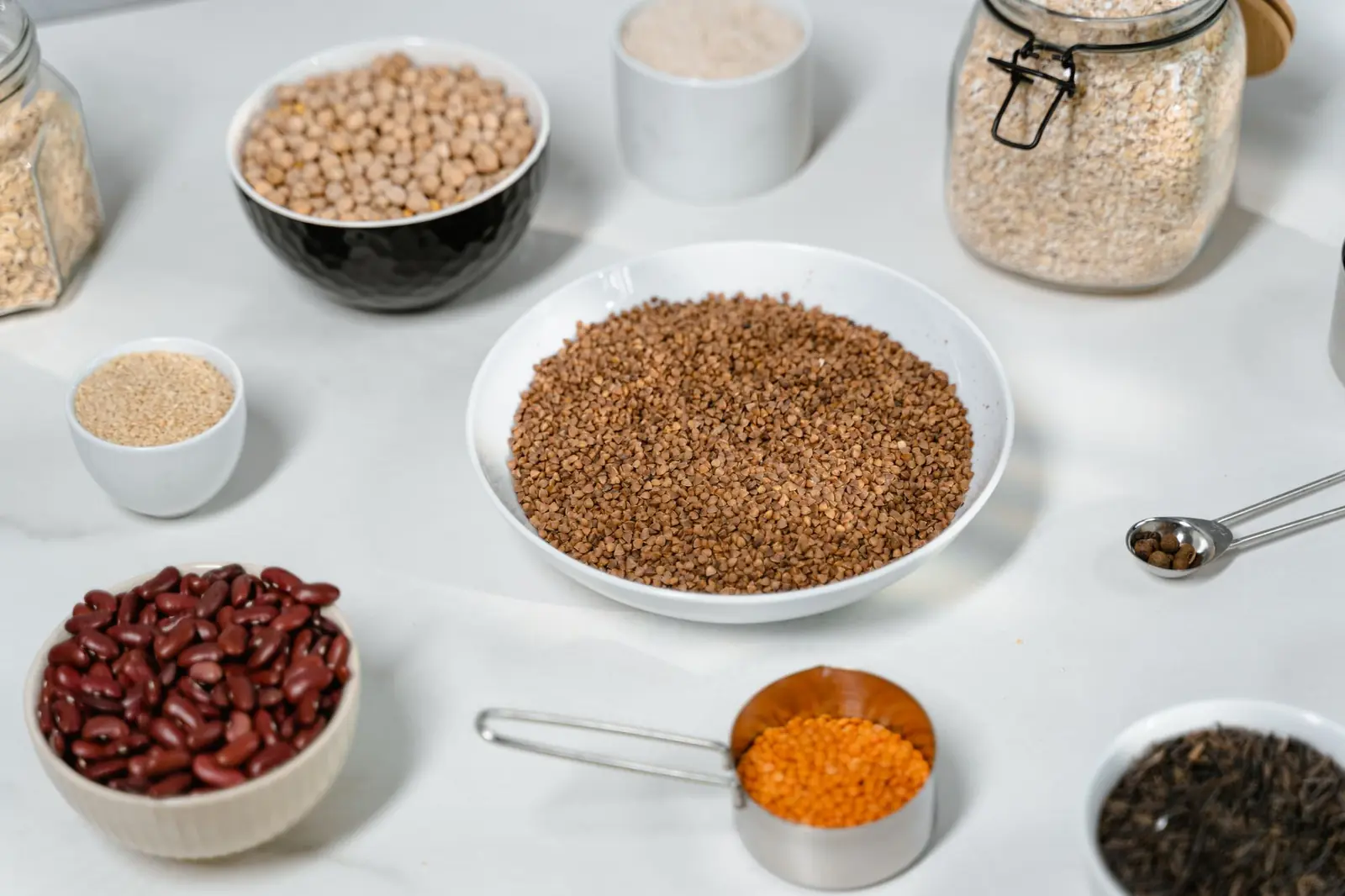Bowls of chickpeas, kidney beans, red lentils, buckwheat and grains with jars of oats on a white table