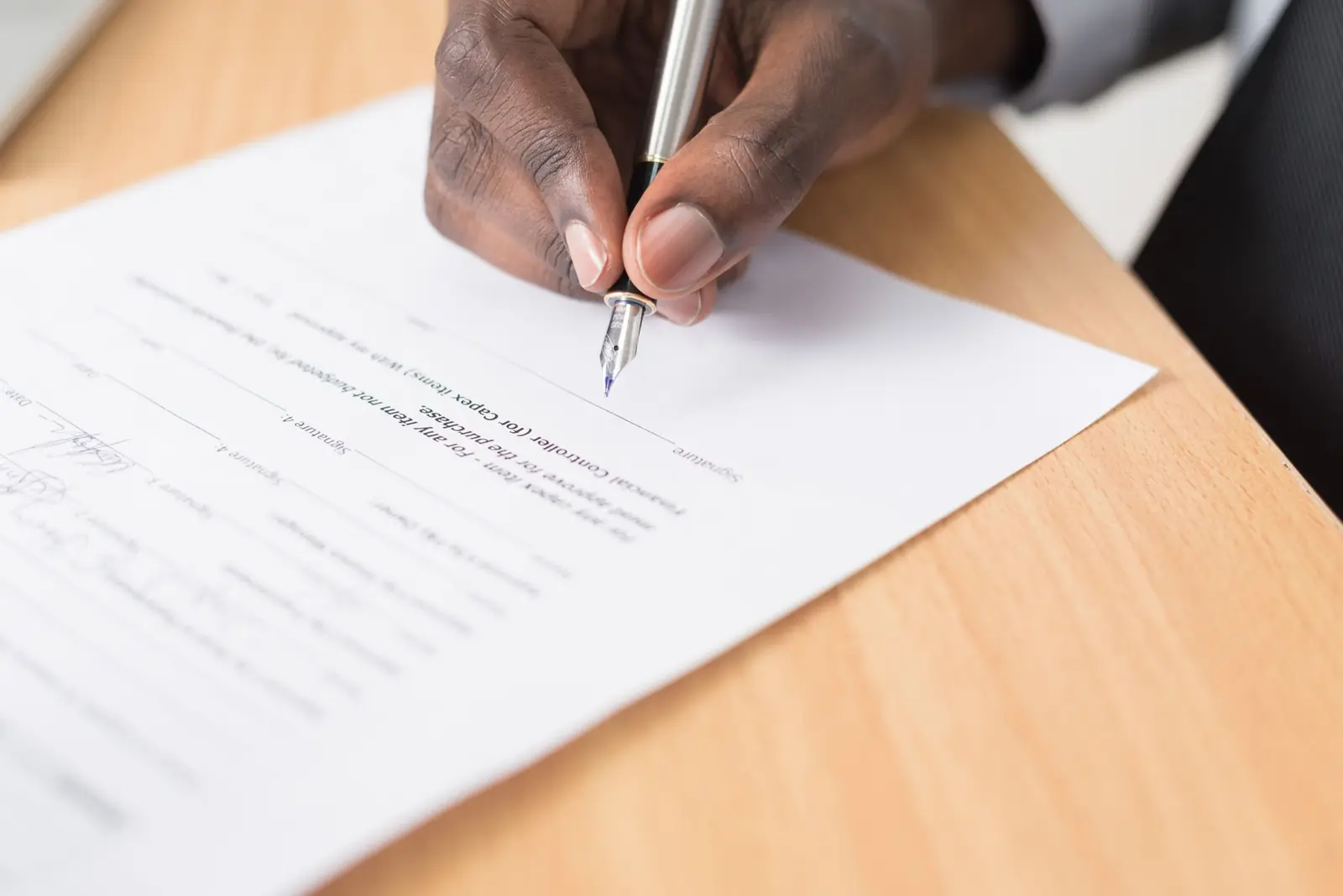 A hand signing a printed document with a silver fountain pen on a light wooden desk
