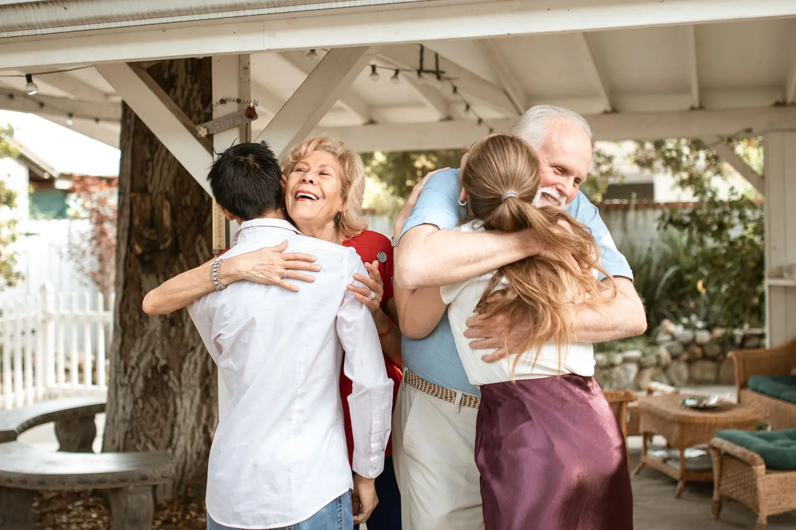 An older couple warmly hugging two younger family members under a backyard pergola