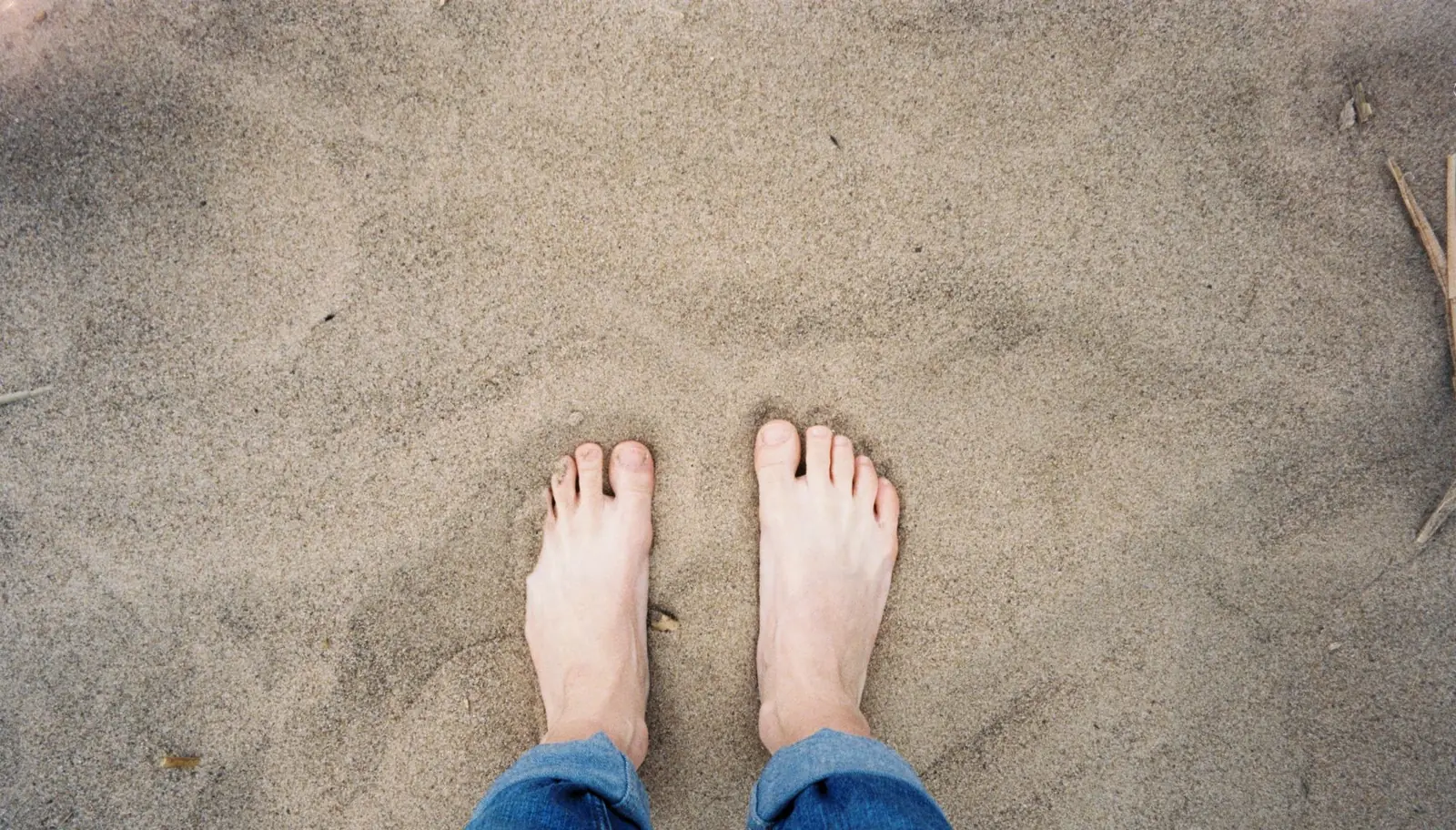 Looking down at a pair of bare feet and rolled-up jeans standing in soft beach sand