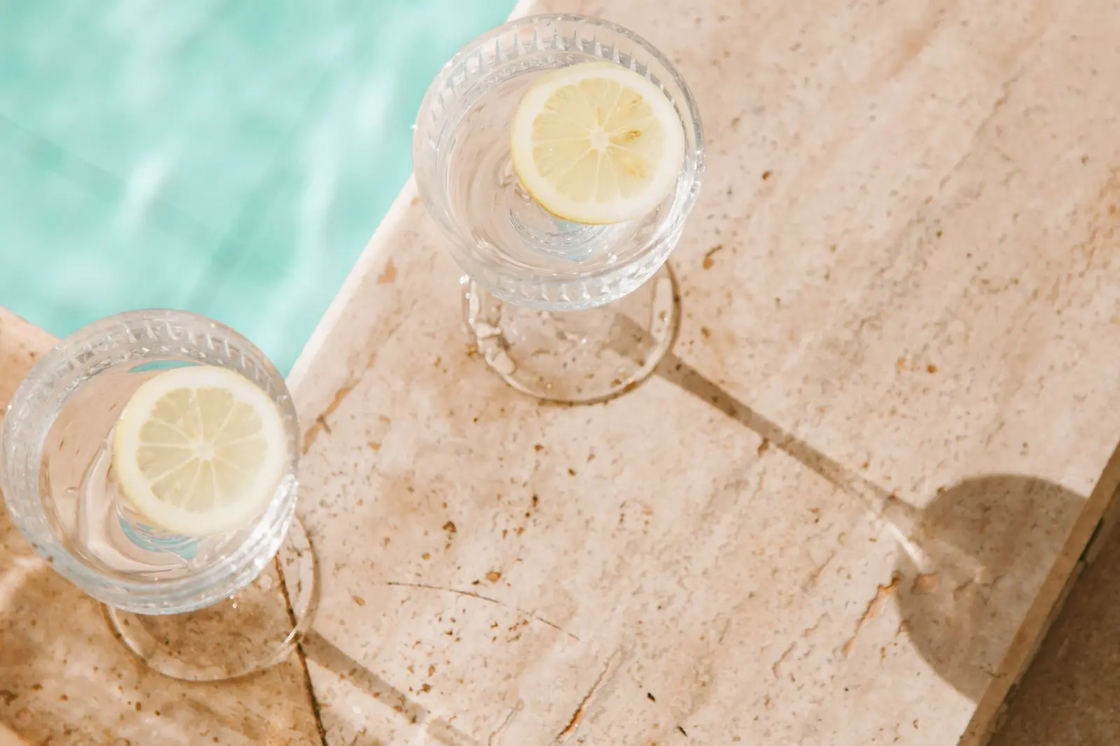 Two glasses of water with lemon slices on sunlit stone beside a swimming pool