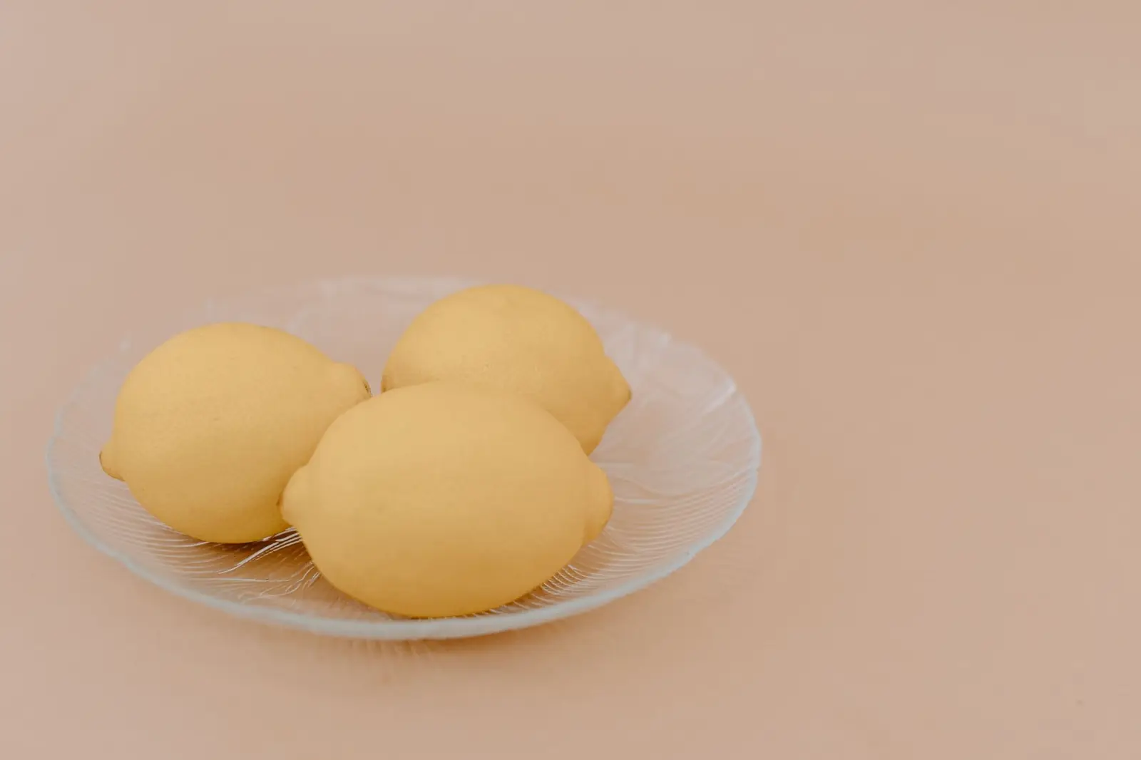 Three lemons on a clear glass plate against a soft peach background