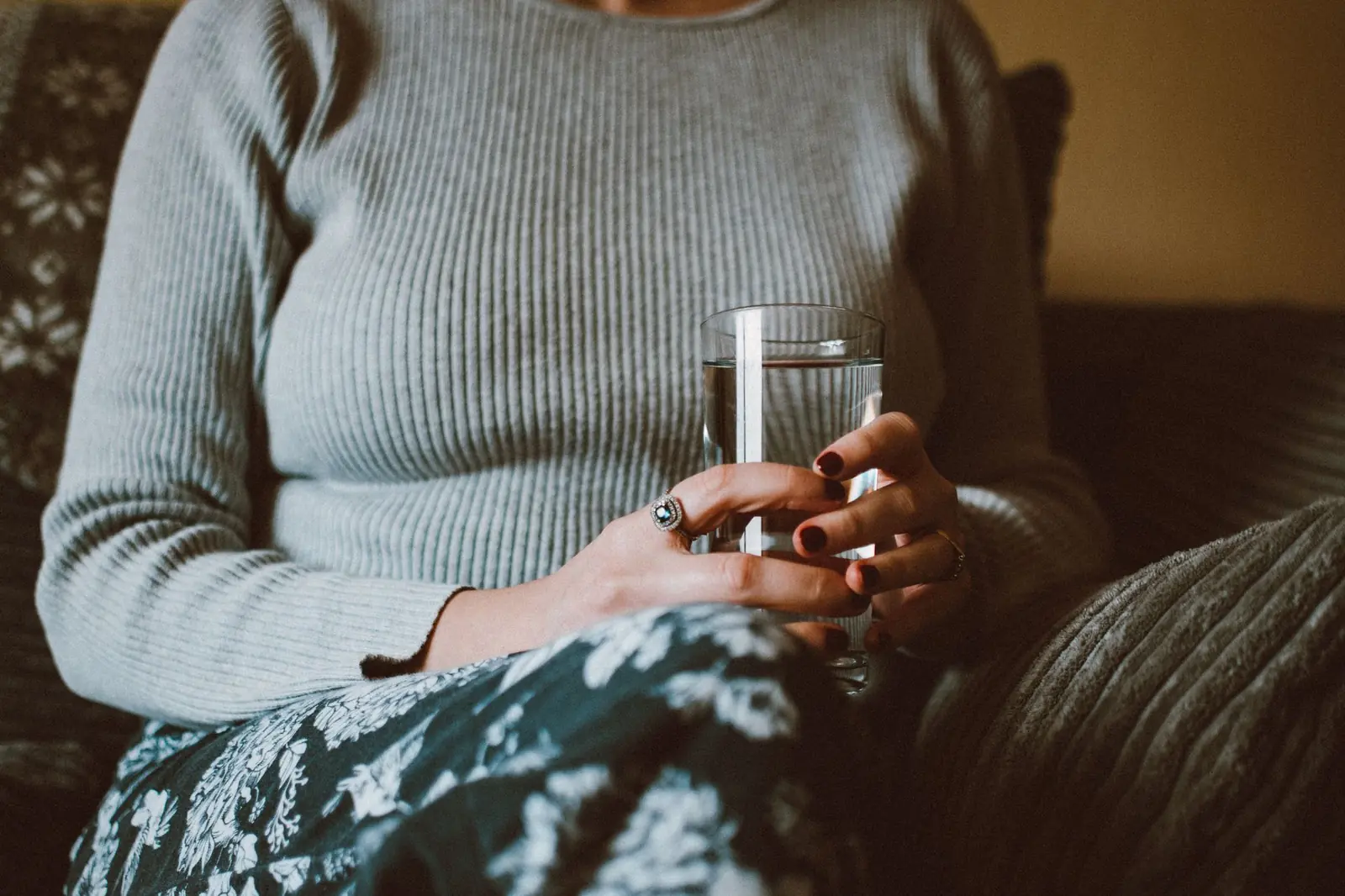 A woman in a grey sweater sitting comfortably and holding a glass of water in both hands