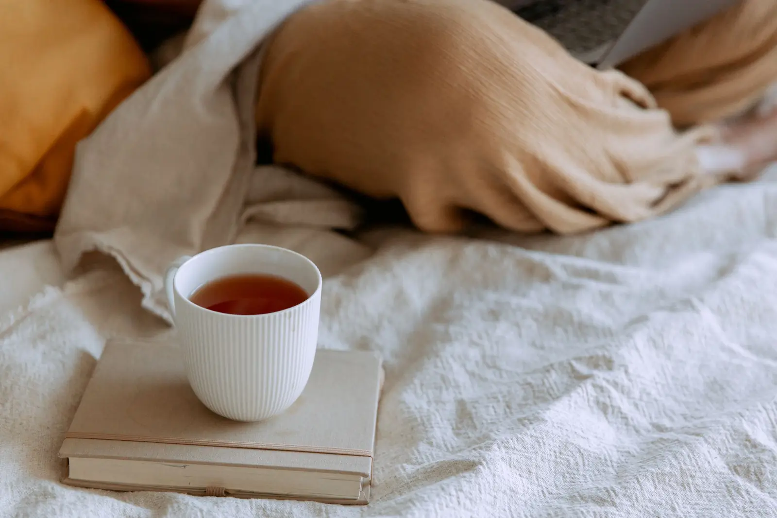 A cup of tea resting on a book on a linen bed.
