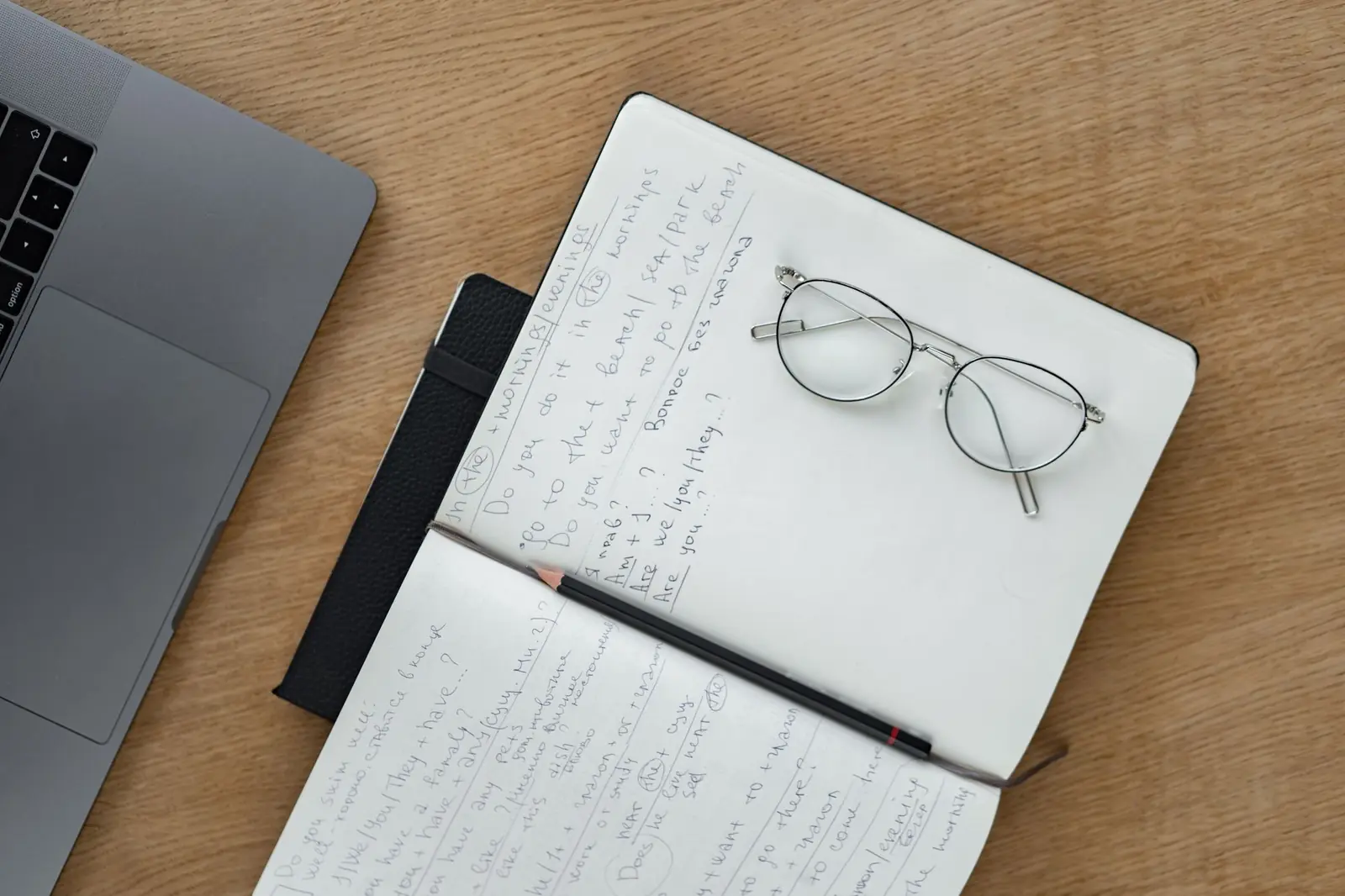 An open notebook with handwritten notes, a pencil and a pair of wire-rimmed glasses on a wooden desk beside a laptop