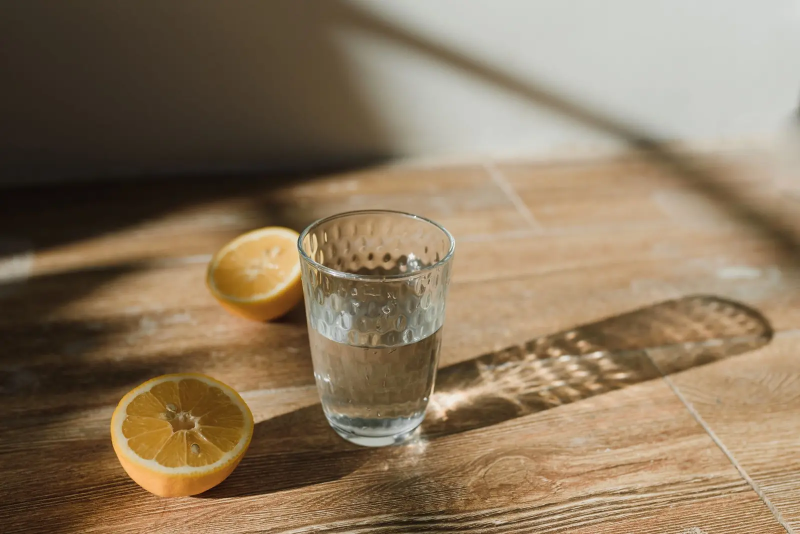 A glass of water and two lemon halves on a wooden table in warm sunlight
