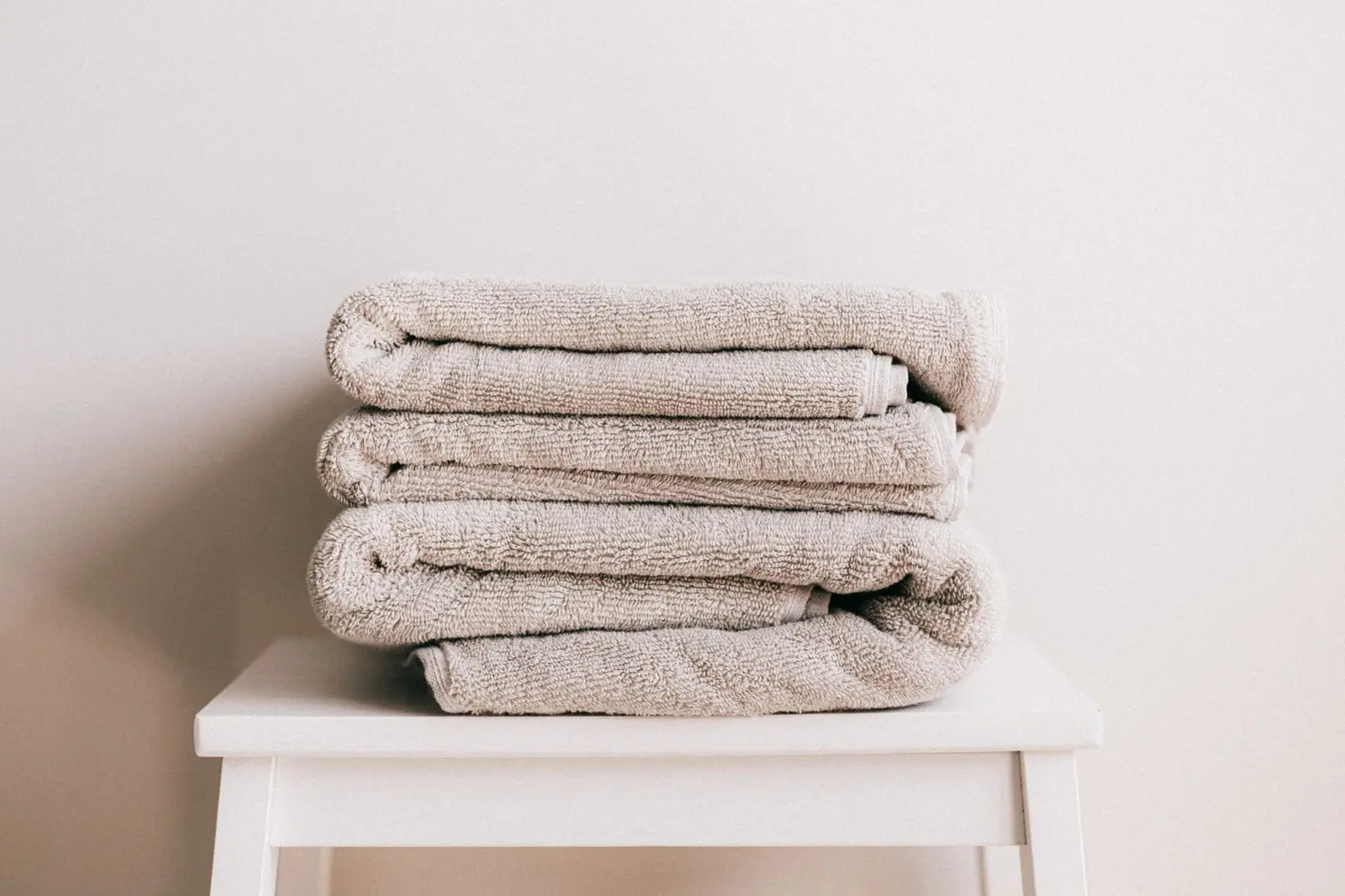 A neat stack of soft beige cotton towels on a white stool against a pale wall