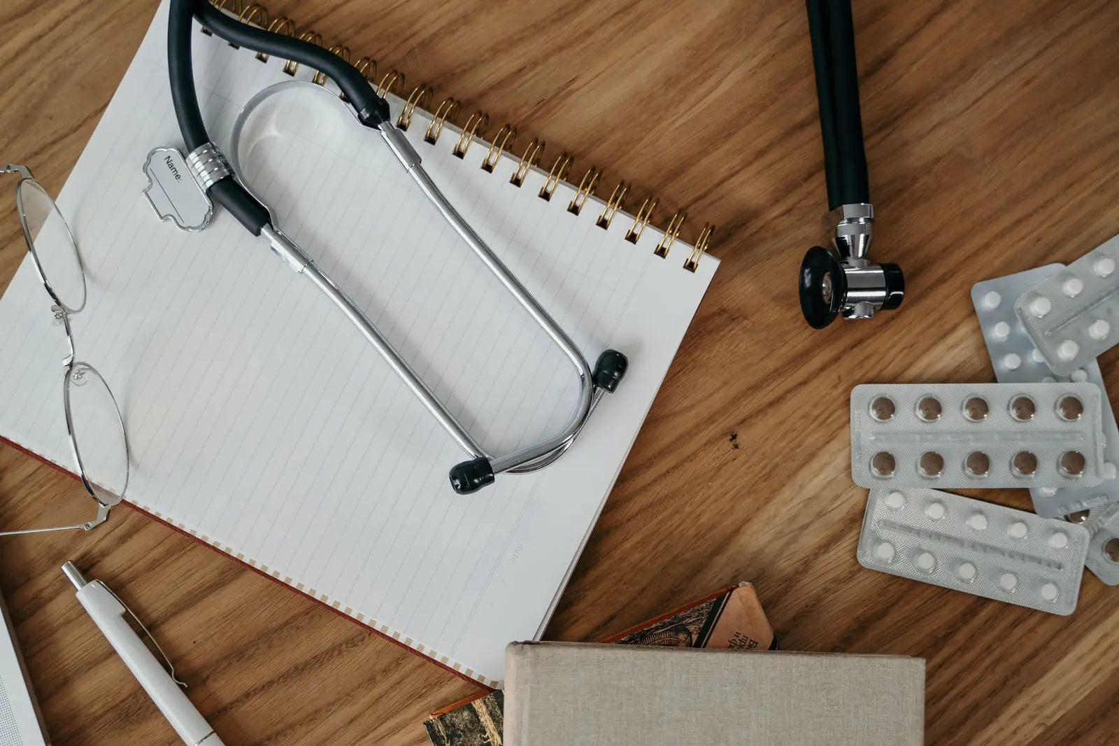 A stethoscope, reading glasses, notebook and blister packs of tablets on a wooden desk