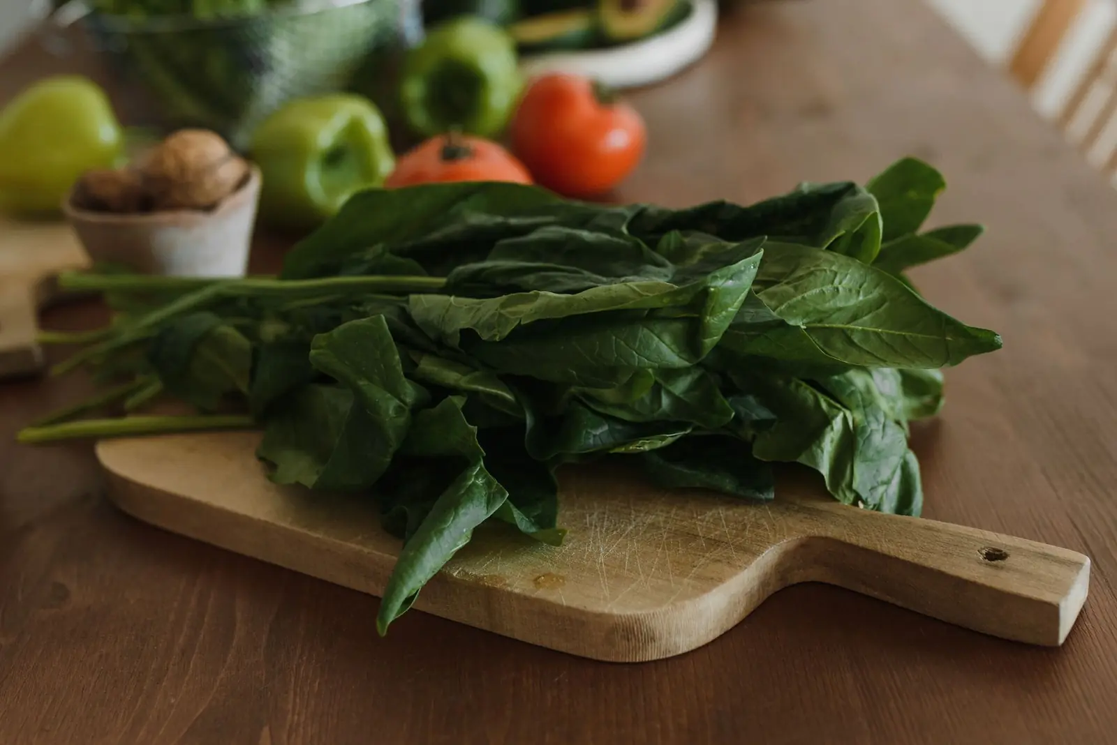 Fresh spinach on a wooden cutting board with peppers and tomatoes on the table behind