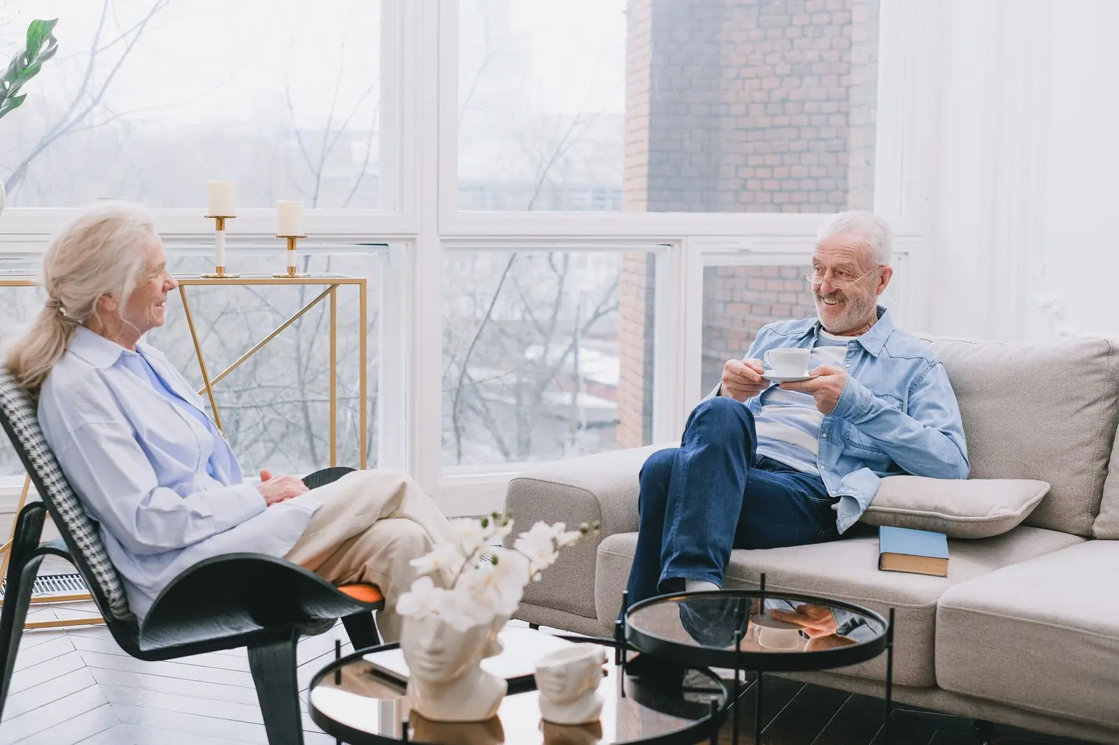 An older couple chatting over cups of tea in a bright living room with tall windows