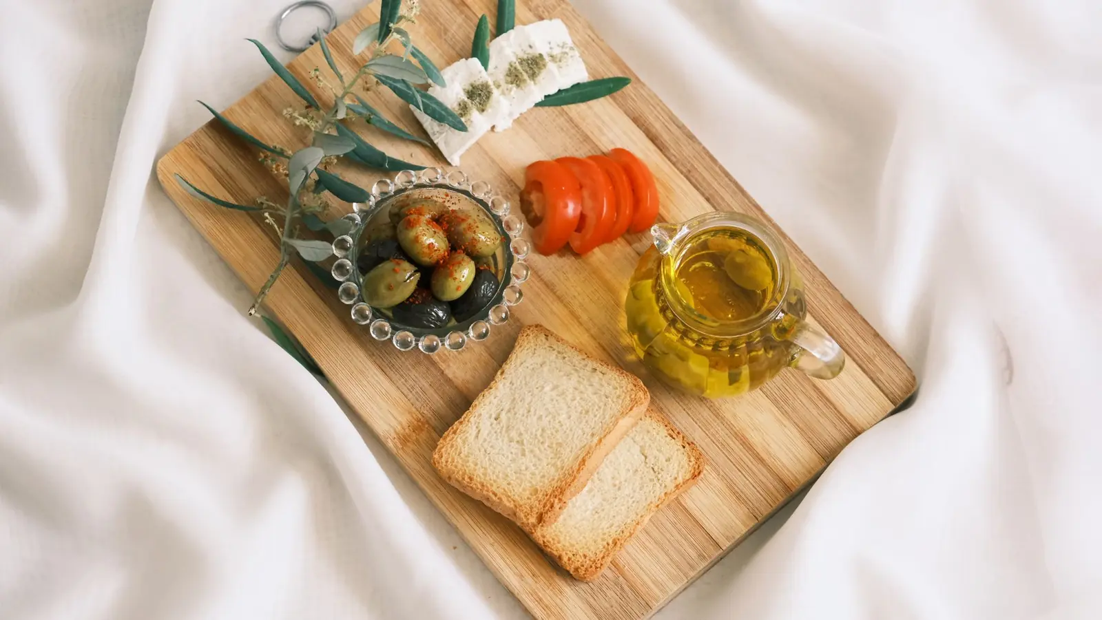 A wooden board with olives, feta, sliced tomato, toasted bread and a small pitcher of olive oil