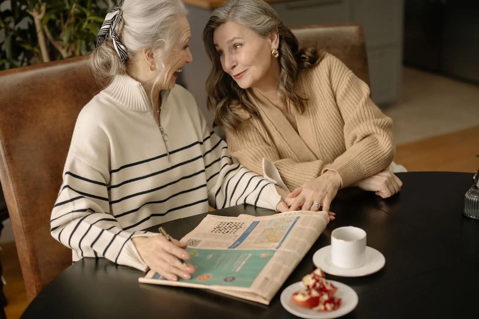 Two older women sharing a laugh over coffee and a newspaper crossword puzzle