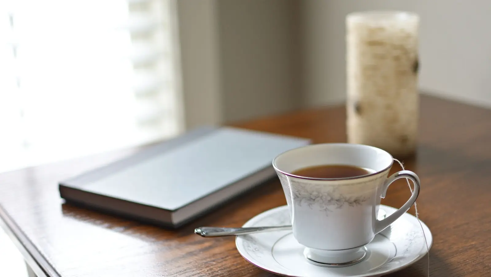 A china cup of tea beside a closed journal on a dark wooden table by a window
