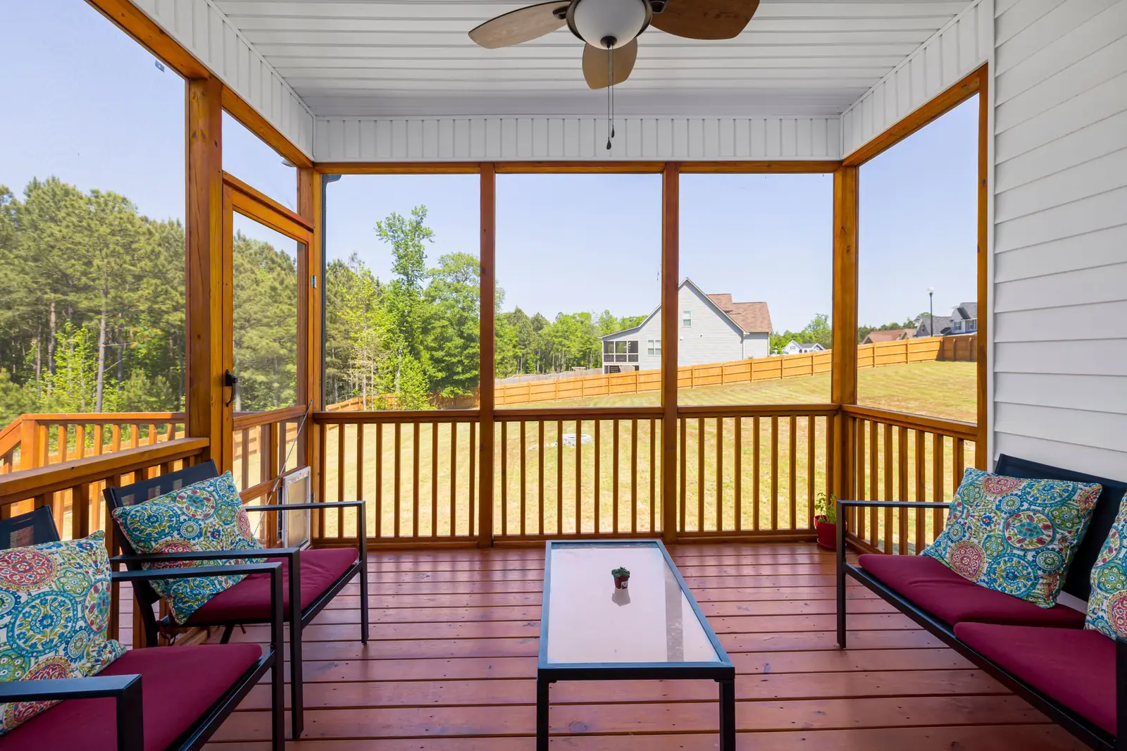 A screened porch with a ceiling fan, cushioned benches and a glass coffee table looking out on a sunny yard