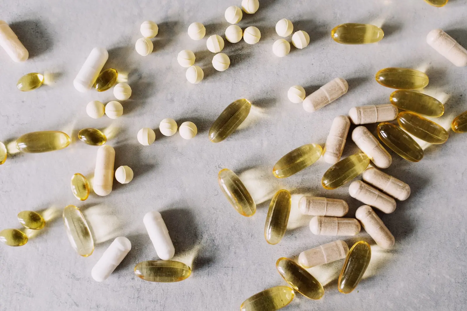 Assorted vitamin capsules, softgels and tablets scattered across a pale stone surface