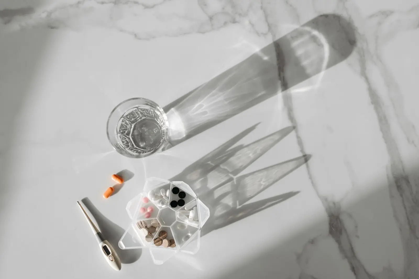 A glass of water, a pill organizer with tablets and a digital thermometer on white marble