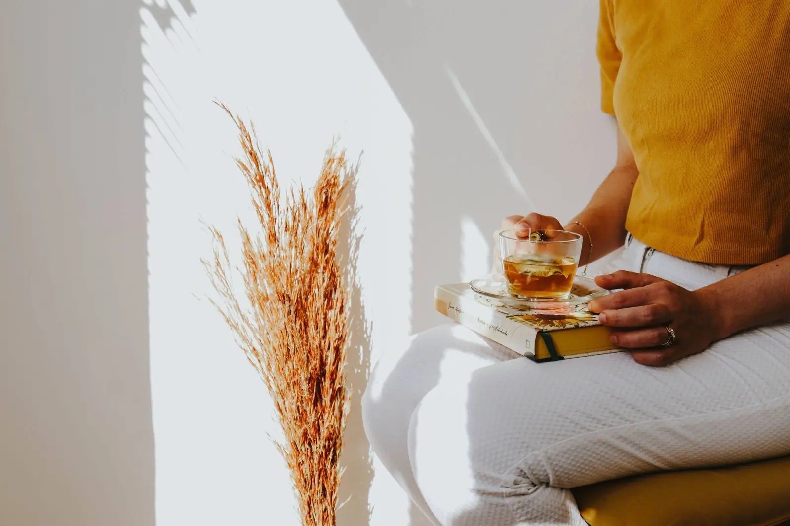 A person resting a glass of tea on a book in their lap beside dried grasses in sunlight