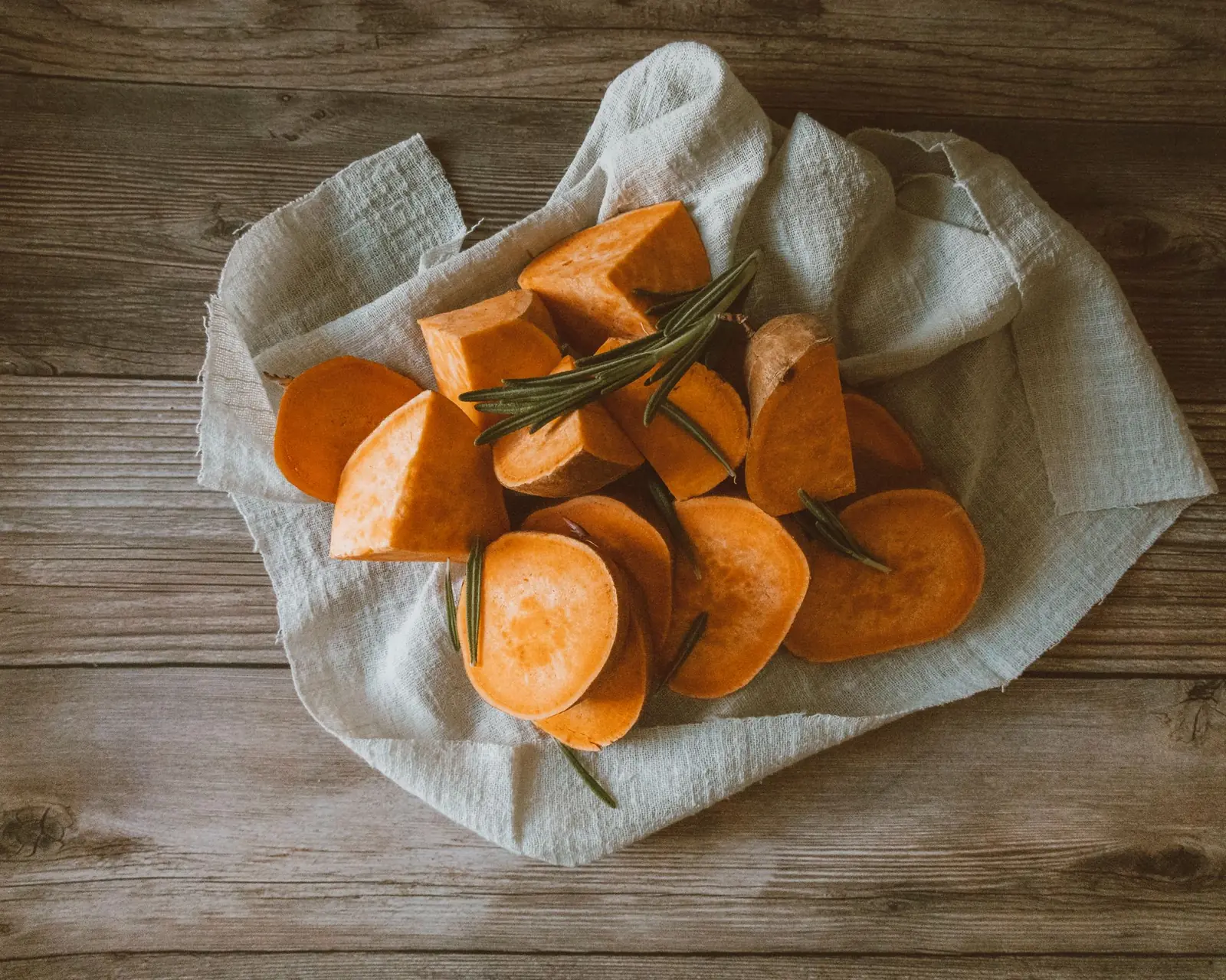 Peeled, chunky slices of orange sweet potato with a rosemary sprig on linen over a wooden table