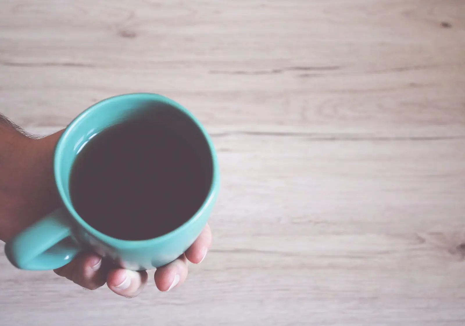 A hand holding a turquoise mug of black coffee over a pale wooden table