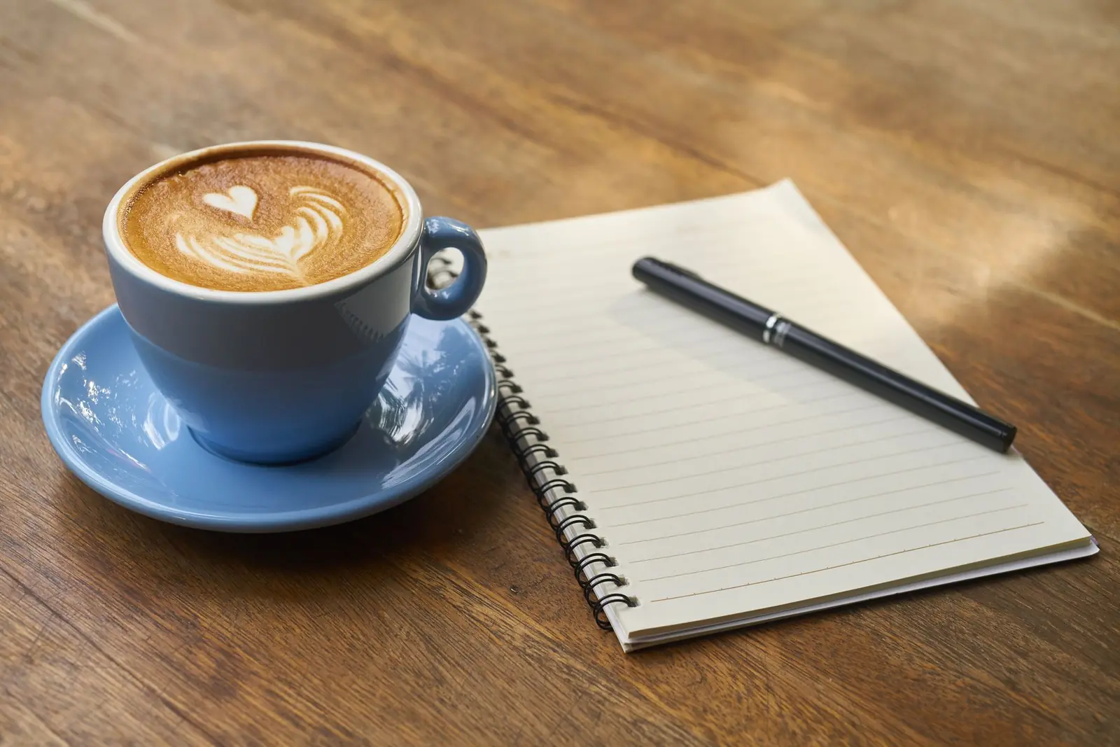A latte in a blue cup beside an open lined notebook and pen on a wooden table