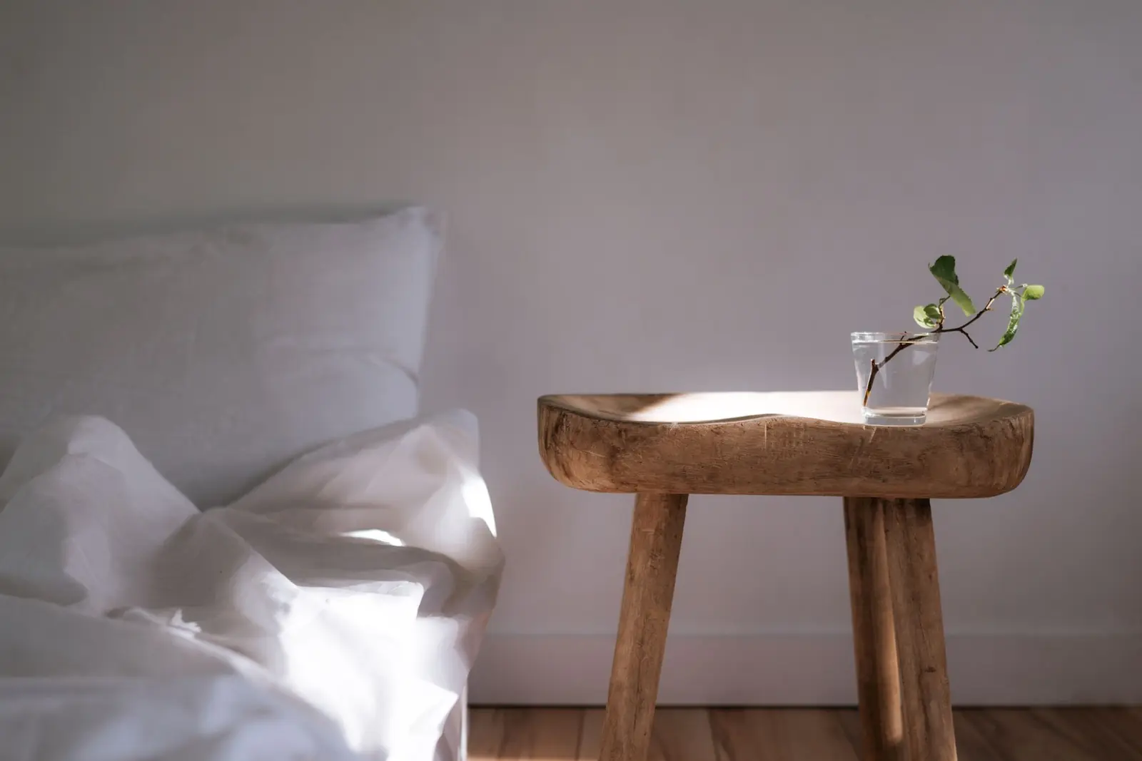 A white bed beside a wooden side table holding a glass of water with a green sprig