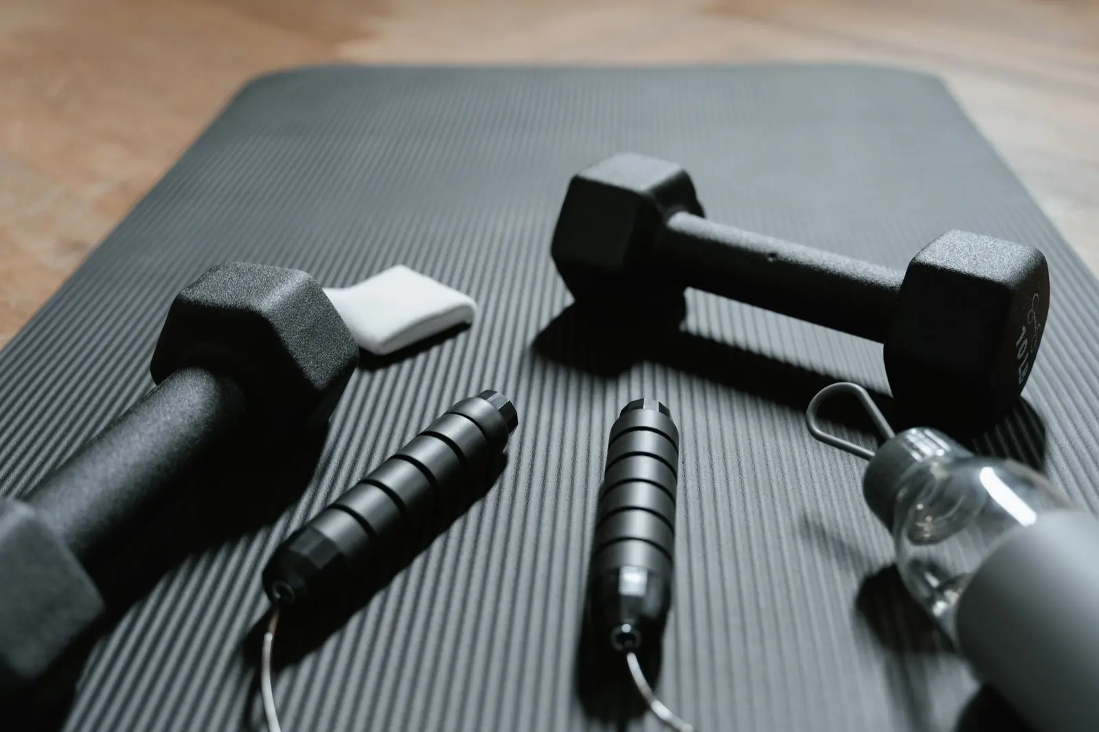 Two black dumbbells, a jump rope and a water bottle resting on an exercise mat on a wooden floor