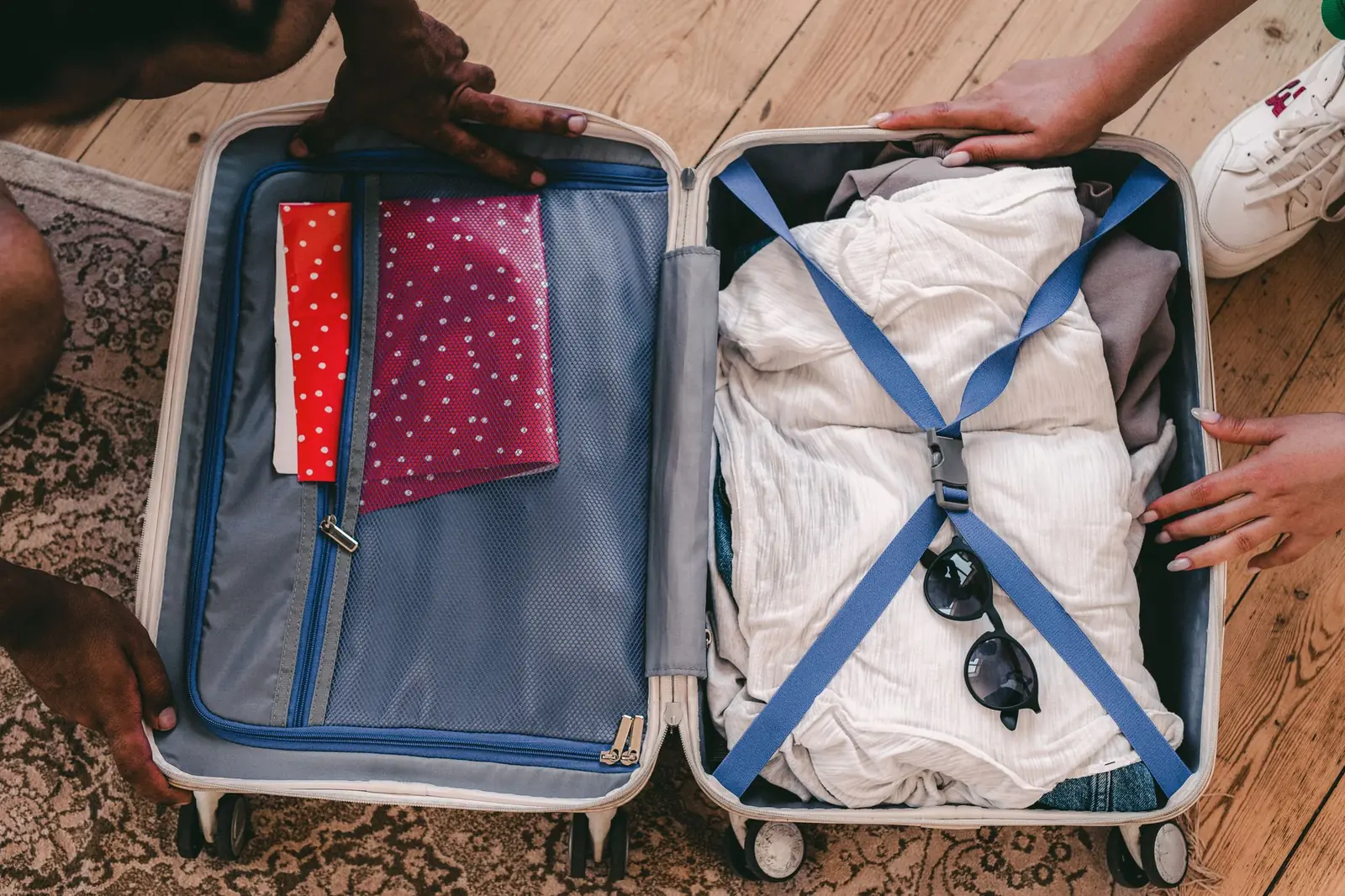 Hands packing an open suitcase with folded white clothes, sunglasses and zipped pouches on a patterned rug