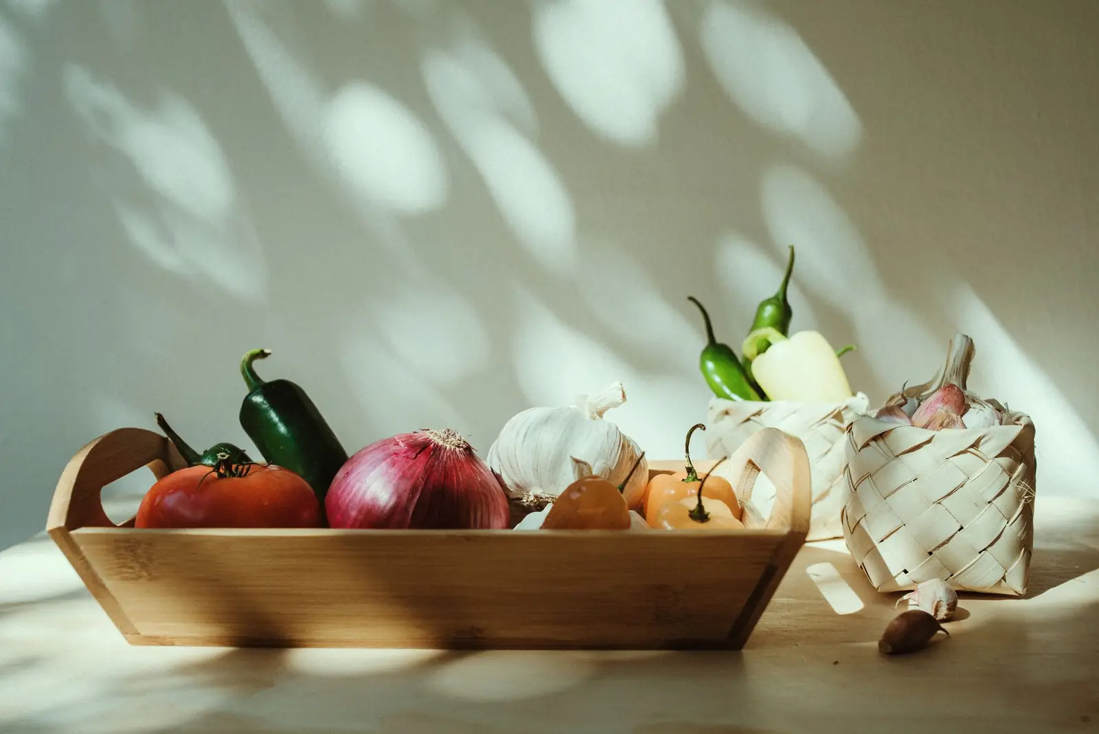 Tomatoes, red onion, garlic and peppers in a wooden tray against a sunlit cream wall
