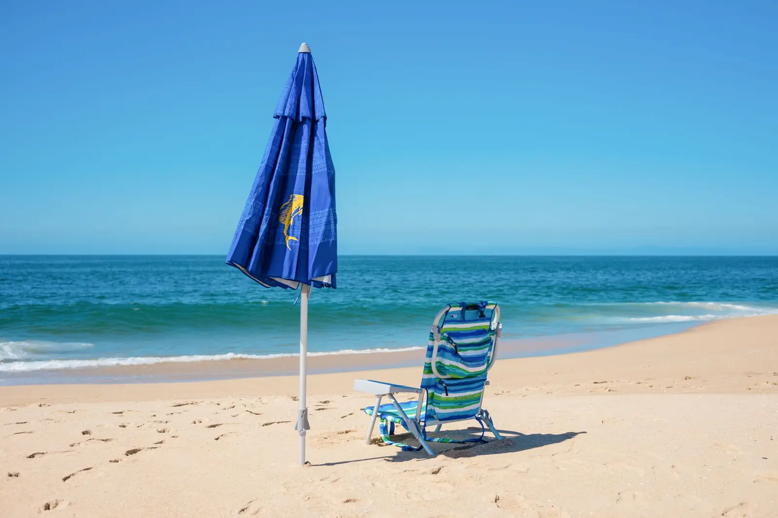 A blue beach umbrella and a striped beach chair on sand by the ocean under a clear sky