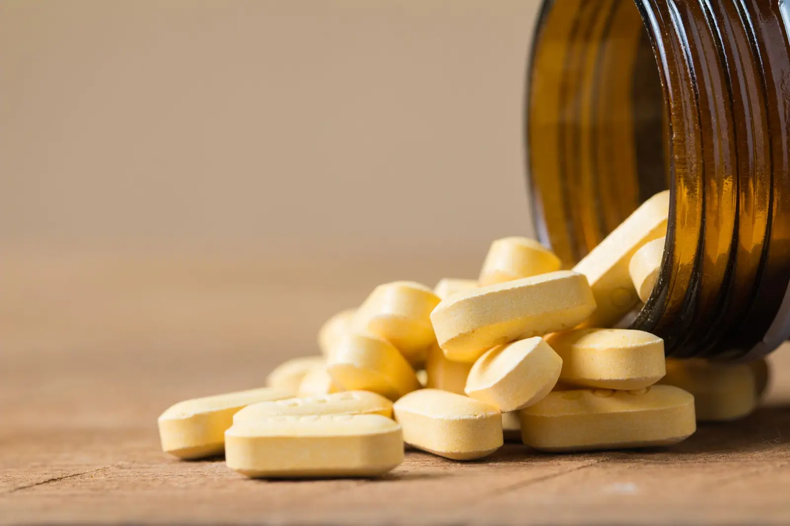 Pale yellow vitamin tablets spilling from an amber glass bottle onto a wooden surface