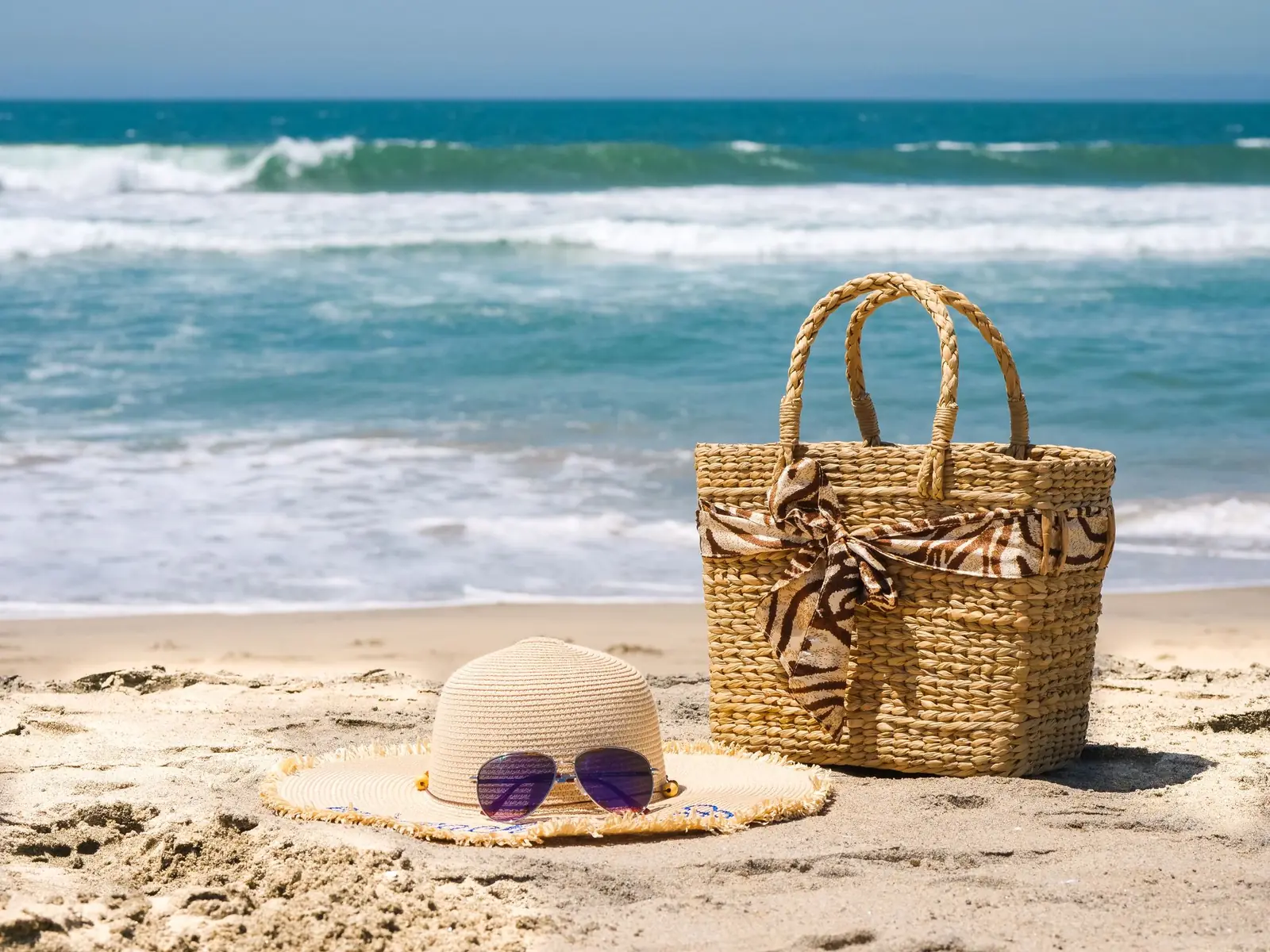 A woven straw beach bag, a sun hat and sunglasses on the sand by the ocean