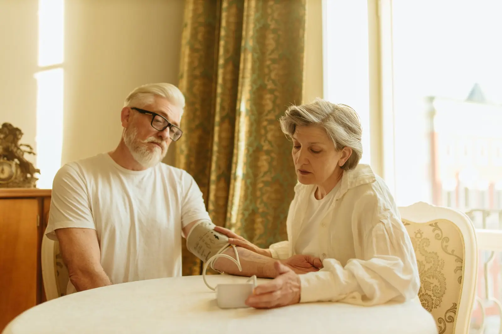An older couple at a sunlit dining table checking blood pressure with a home cuff and digital monitor.