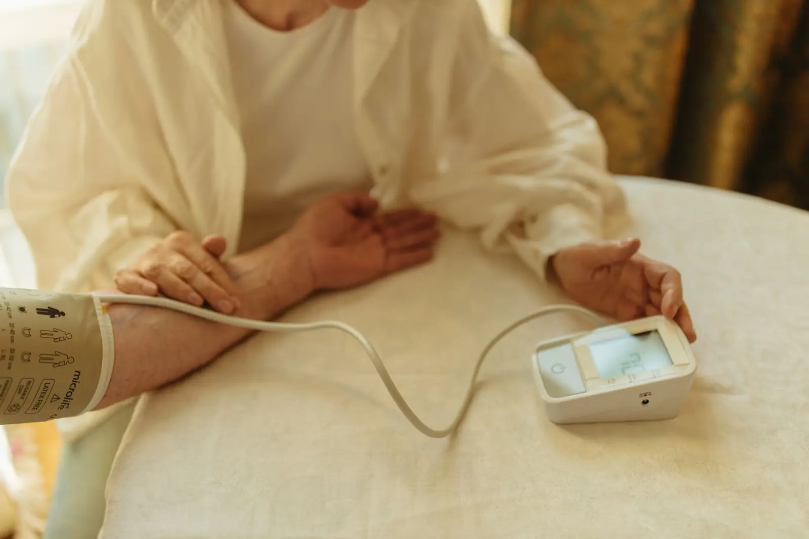 A home blood pressure monitor on a cream tablecloth beside a resting arm in soft light.