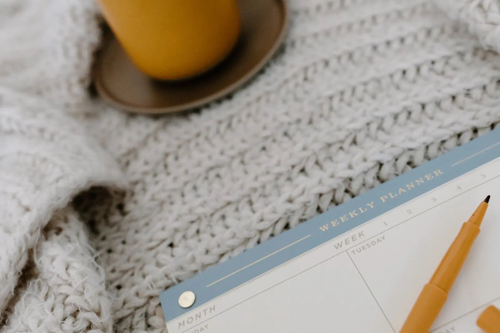 A weekly planner and yellow pen beside a warm drink on a cream knitted blanket.