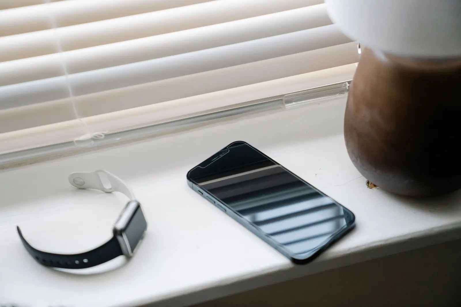 A smartphone and wristwatch resting on a sunlit white windowsill beside window blinds.