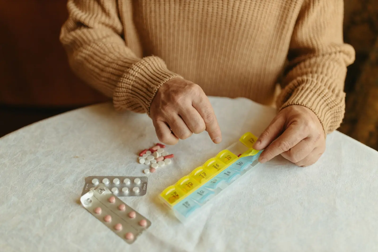 Hands in a warm knit sweater sorting tablets into a weekly pill organizer on a white tablecloth.