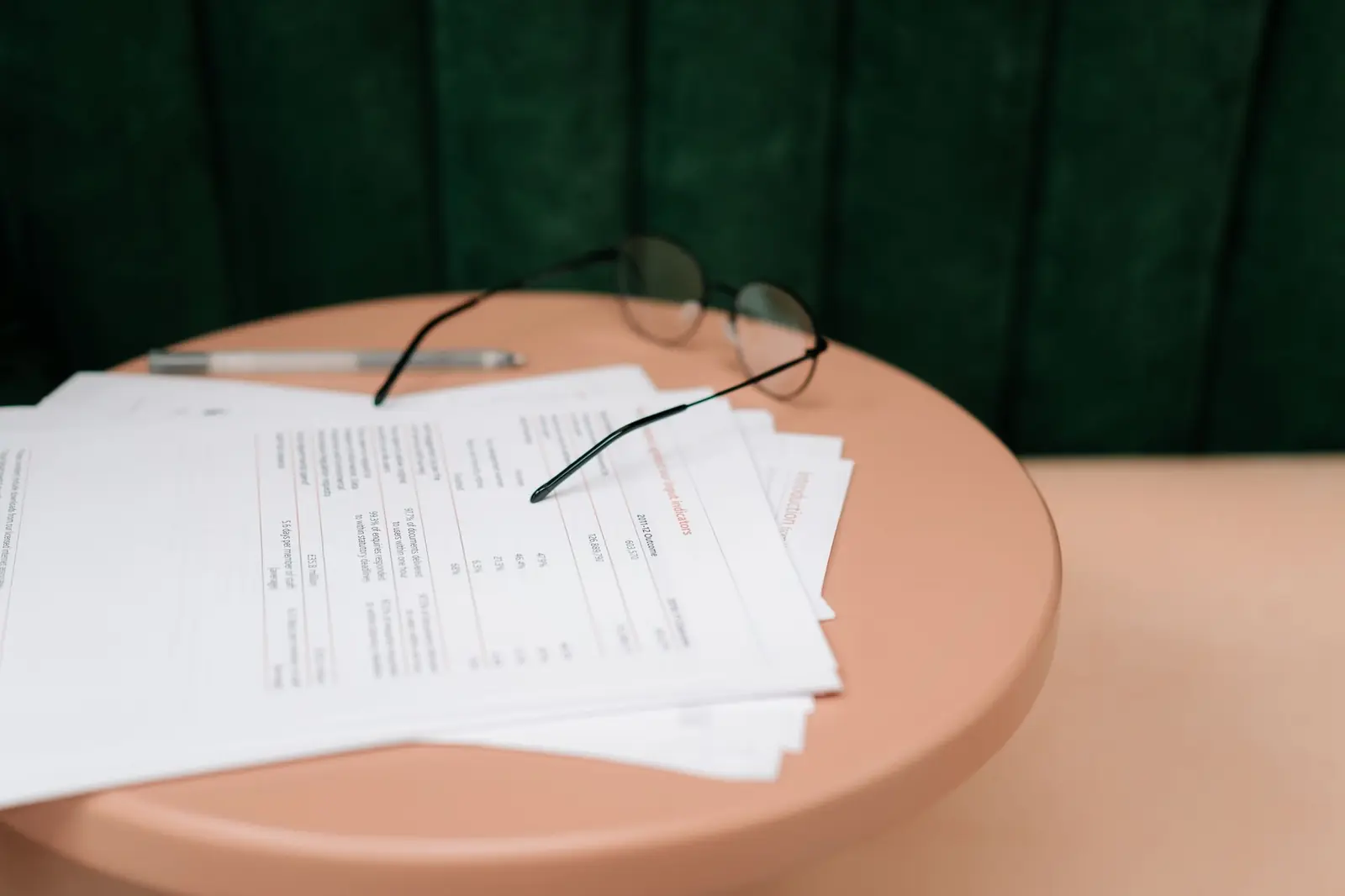 A stack of printed forms with reading glasses and a pen on a peach-colored side table.