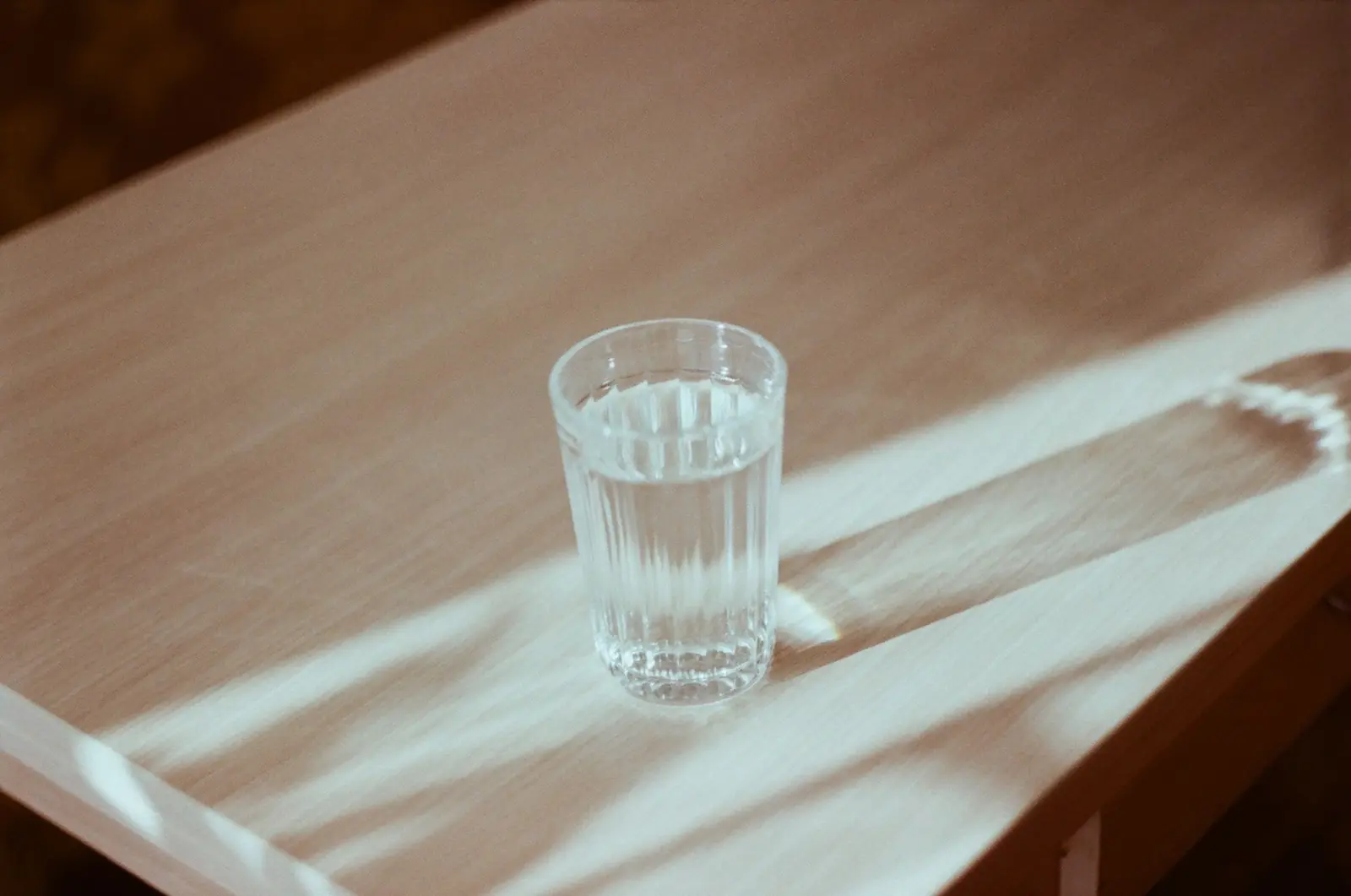 A clear glass of water on a wooden table, crossed by soft bands of afternoon sunlight.
