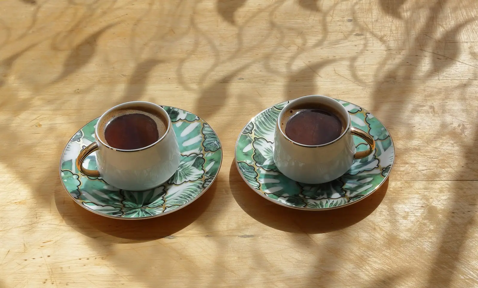 Two cups of coffee on green leaf-patterned saucers on a wooden table dappled with sunlight.